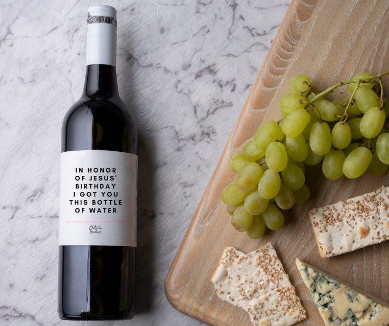 May include: A bottle of red wine with a white label that reads "In honour of Jesus' birthday I got you this bottle of water". The bottle is on a white marble surface with a wooden chopping board in the background. The chopping board has a bunch of green grapes, a wedge of blue cheese, and two pieces of flatbread with sesame seeds.