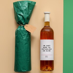 May include: A bottle of amber-colored liquid with a white label that reads "IM JUST HERE TO ESTABLISH AN ALIBI". The bottle is next to a wrapped bottle in green paper with a blank tag.