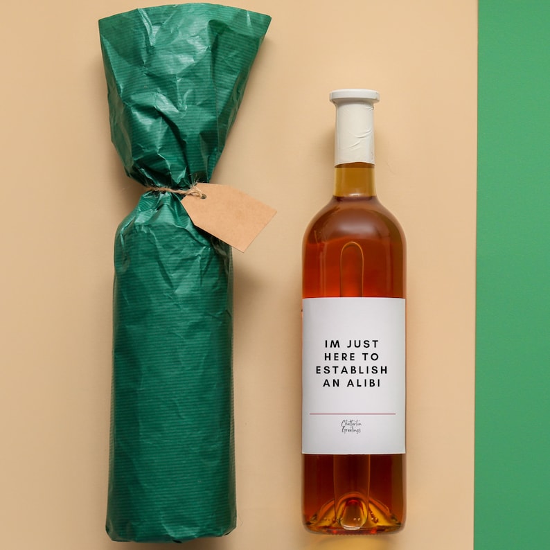 May include: A bottle of amber-colored liquid with a white label that reads "IM JUST HERE TO ESTABLISH AN ALIBI". The bottle is next to a wrapped bottle in green paper with a blank tag.