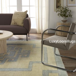 Pode incluir: Um tapete bege claro com um padr&atilde;o geom&eacute;trico azul. O tapete est&aacute; em uma sala de estar com um sof&aacute; marrom, uma cadeira cromada e uma mesa auxiliar de madeira.