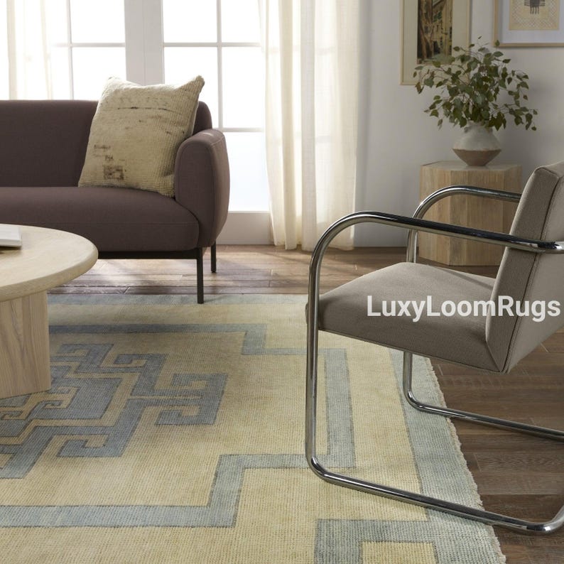 Pode incluir: Um tapete bege claro com um padr&atilde;o geom&eacute;trico azul. O tapete est&aacute; em uma sala de estar com um sof&aacute; marrom, uma cadeira cromada e uma mesa auxiliar de madeira.
