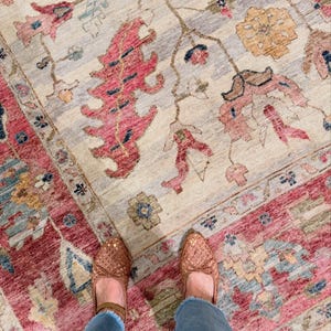 May include: An overhead shot of a beige and red patterned rug with floral and geometric designs. The rug has a distressed look with a border of red and blue. Woven brown shoes and blue jeans are visible.