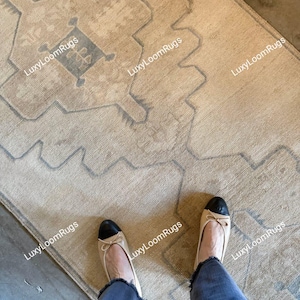 May include: Beige and gray patterned rug with geometric designs. The rug has a central medallion and border patterns. The image also shows a person wearing beige ballet flats with black toes and blue jeans.