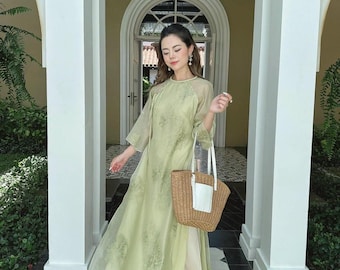 Pode incluir: Uma mulher vestindo um vestido floral verde claro com uma sobreposição transparente e uma bolsa de palha branca e marrom. Ela está em pé em uma porta com colunas brancas de cada lado.