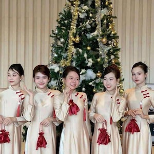 May include: Five people wearing cream-colored dresses with long sleeves and mandarin collars. Each person holds a small red pouch with a tassel. A decorated Christmas tree is in the background.