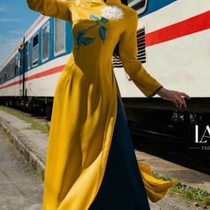 May include: A woman in a yellow and blue traditional Vietnamese dress, called an Ao Dai, stands in front of a train. She is holding a fan and has a flower embroidered on her dress.