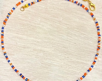 May include: A beaded necklace featuring a pattern of orange, blue, and white beads. The necklace has gold-colored clasps and connectors. The beads are small and evenly spaced, creating a delicate and colorful accessory.