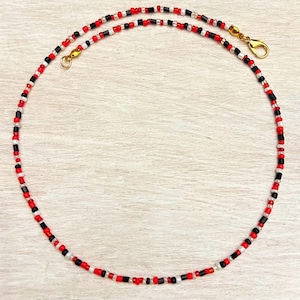 Puede incluir: Un collar de cuentas rojas, negras y blancas. Tiene un cierre dorado. Las cuentas son pequeñas y están espaciadas uniformemente, creando un accesorio delicado y colorido. El collar se muestra sobre un fondo de madera clara.