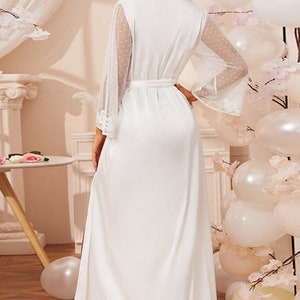 May include: A white satin robe with sheer, polka-dot sleeves and lace cuffs. The robe has a matching belt and a high collar. The long, flowing robe is suitable for bridal wear or a special occasion. The background includes balloons and floral decorations.