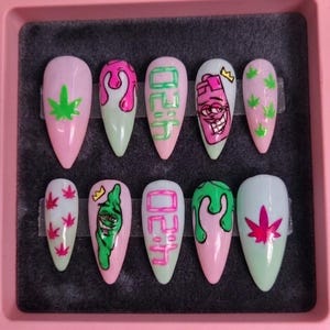 Op de afbeelding: Een set van tien kunstnagels in een roze etui. Elke nagel heeft een uniek ontwerp, waaronder cannabisbladeren, druipende verf, cartoonfiguren en het woord "lit" in groen en roze. De nagels hebben een glanzende afwerking.