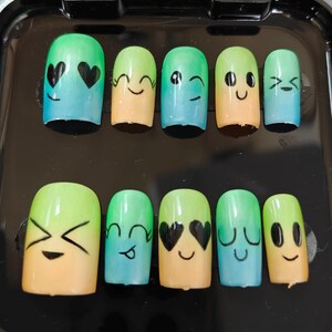 May include: A set of ten fake nails with a green and blue ombre design. Each nail has a different emoji face painted on it, including hearts, winking eyes, and a smiley face.