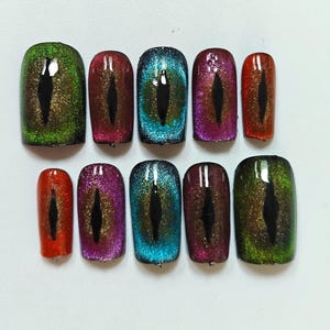 Könnte beinhalten: Ein Set aus zehn künstlichen Fingernägeln mit magnetischem Katzenaugen-Effekt in verschiedenen Farben wie Grün, Rot, Blau und Violett. Jeder Nagel ist mit einem zentralen, vertikalen schwarzen Schlitz versehen, der wie eine Pupille wirkt.