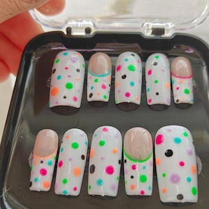 May include: A set of ten false nails with a white base and colorful polka dots. The nails are in a black plastic case with a clear lid.