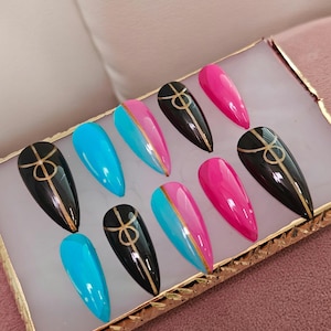 May include: A set of almond-shaped press-on nails in various colors. The set includes black nails with gold designs, bright pink nails, and nails split between turquoise and pink with a gold stripe. The nails are displayed on a white surface with a gold border.