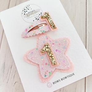 May include: Two pink glitter hair clips, one star-shaped and one barrette, each with a gold glitter "1". The clips are presented on a white card with the text "BOWS BOUTIQUE".