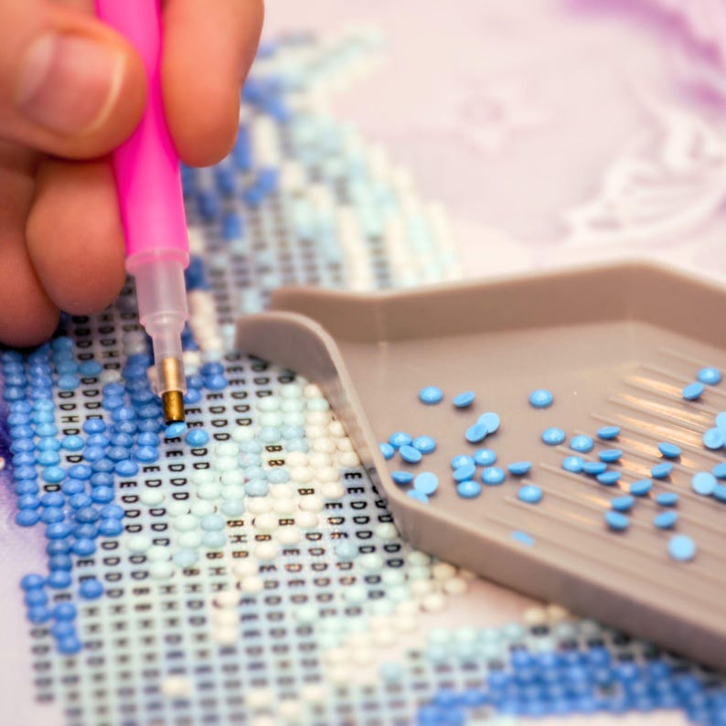 May include: A close-up of a diamond painting project in progress. A hand holds a pink stylus, placing a small, light blue diamond onto the canvas. A gray tray holds additional blue diamonds, and the canvas features a pattern of blue and white squares.