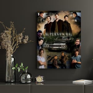 May include: A framed poster featuring a collage of images of actors and a black car. The word "SUPERNATURAL" is printed in white capital letters across the center of the poster. The poster is displayed on a dark gray wall.