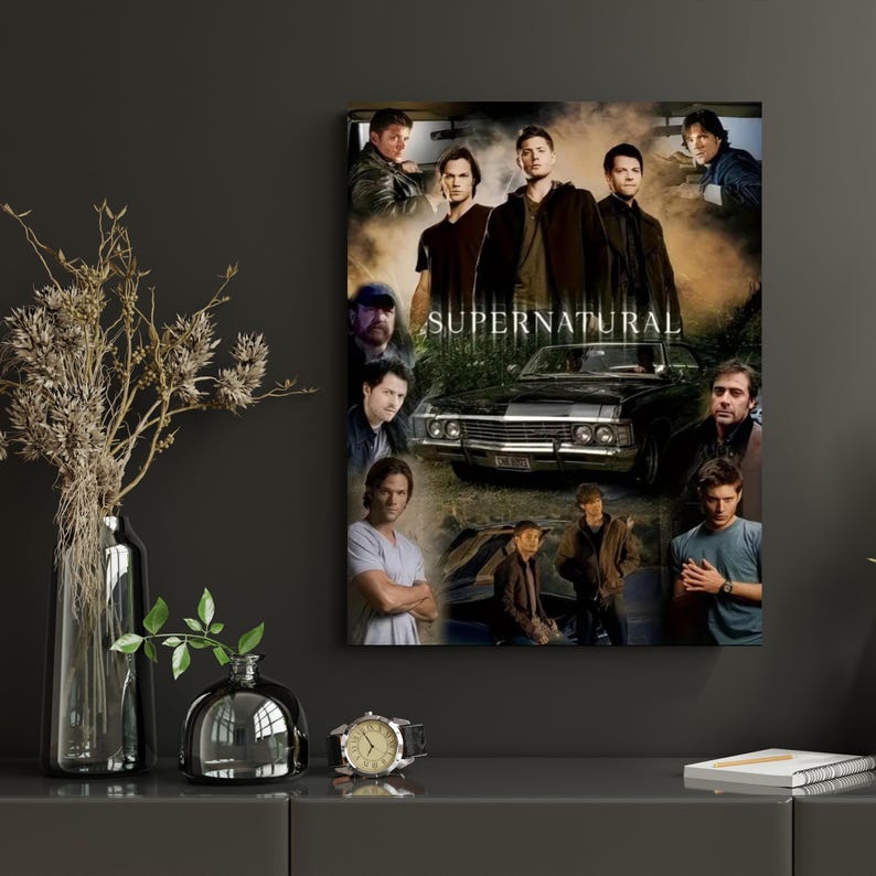 May include: A framed poster featuring a collage of images of actors and a black car. The word "SUPERNATURAL" is printed in white capital letters across the center of the poster. The poster is displayed on a dark gray wall.