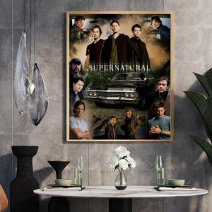 May include: A framed poster featuring multiple images of actors from the television show "Supernatural". The poster includes the show's title and a black car. The poster is displayed on a gray wall, with a table and decorative items in front.