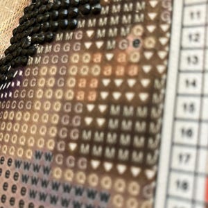 May include: Close-up of a diamond painting in progress. The image shows a section of the canvas with black diamond-shaped beads and a grid of symbols and letters. The color scheme is a mix of brown, white, and black.