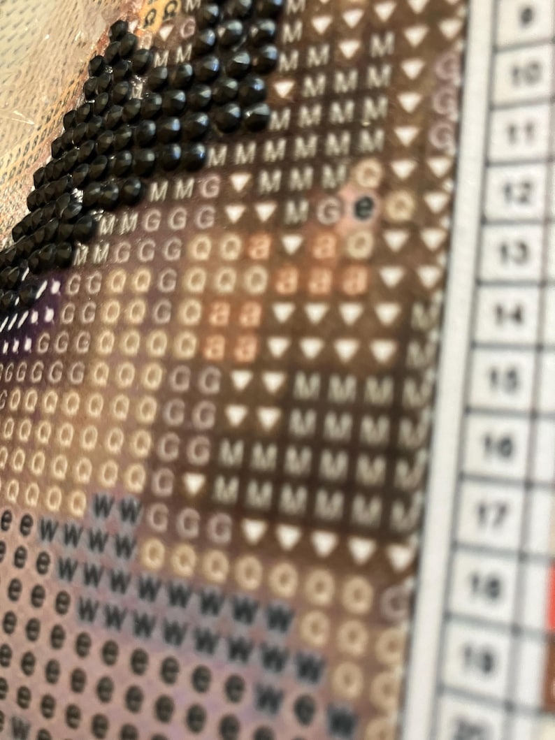 May include: Close-up of a diamond painting in progress. The image shows a section of the canvas with black diamond-shaped beads and a grid of symbols and letters. The color scheme is a mix of brown, white, and black.