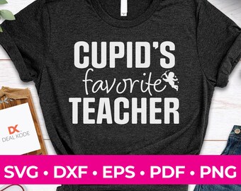 以下が含まれることがあります： 暗いグレーのTシャツに、「CUPID'S favorite TEACHER」という白い文字が書かれています。 「favorite」の隣には、矢を持ったキューピッドの小さな白い画像があります。