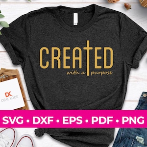 Puede incluir: Una camiseta gris oscuro con las palabras "CREATED with a purpose" en letras doradas. Una cruz dorada está por encima de la palabra "CREATED".