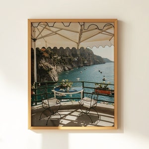 May include: A framed photograph of a table and two chairs on a balcony overlooking a blue sea with a white awning above. The balcony is surrounded by a metal railing and there are green trees and cliffs in the background.