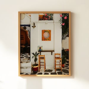 May include: A framed photograph of a white building with a wooden door and two chairs in front of it. The building has a sign that says "Mandala" and a flower pot with green plants.