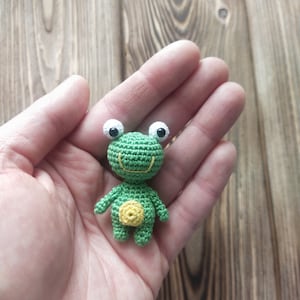 May include: A small, handmade crochet frog in shades of green and yellow. The frog has large white eyes with black pupils, a yellow belly, and a smiling mouth. The frog is held in a hand against a wooden background.