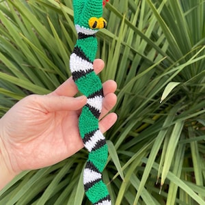 May include: A crocheted snake toy in green, black, and white. The snake's head is green with yellow eyes and a red tongue. The body features a repeating pattern of green, black, and white stripes. The toy is held against a backdrop of green foliage.