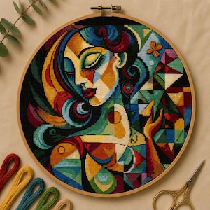 May include: A circular wooden embroidery hoop containing a cross-stitch pattern of a stylized face with closed eyes. The design features vibrant, geometric blocks of color including blue, green, yellow, and red, with a dark background.