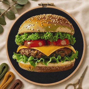 May include: A cross-stitch embroidery project in a round wooden hoop featuring a detailed hamburger with a golden-brown bun, green lettuce, red tomato slices, yellow cheese, and a brown patty, all stitched against a solid black fabric background.