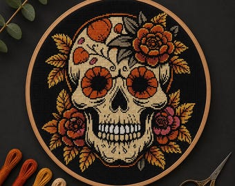 Könnte beinhalten: Eine Kreuzstichstickerei in einem runden Holzrahmen mit einem beigen Totenkopf, umgeben von orangefarbenen und rosafarbenen Blumen und Blättern auf schwarzem Stoff, begleitet von mehreren Stickgarnsträngen und einer Metallschere.