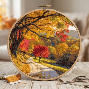 May include: A circular wooden embroidery hoop containing a cross-stitch pattern of an autumn landscape with a winding path, vibrant yellow and red trees, and dark tree branches. Beside the hoop are yellow embroidery floss and metal scissors.