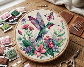 May include: A circular wooden embroidery hoop containing a cross-stitch pattern of a hummingbird in flight, surrounded by pink and purple flowers and a small butterfly. The design is stitched on light beige fabric, featuring vibrant green and pink hues.