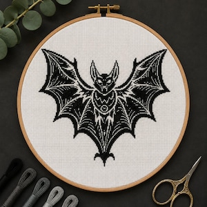 May include: A circular wooden embroidery hoop containing white fabric with a black cross-stitch design of a bat with its wings spread wide. The bat features detailed line work and a small circular emblem on its chest, centered within the hoop.
