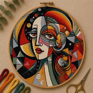 May include: A circular embroidery hoop containing a cross-stitch portrait of a face with geometric, cubist-inspired features. The design uses a vibrant palette of red, orange, yellow, blue, and black thread, creating a complex, segmented mosaic pattern.