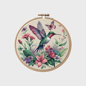 May include: A circular wooden embroidery hoop containing a cross-stitch design on cream-coloured fabric. The embroidery features a central hummingbird with green, purple, and red plumage, surrounded by pink and purple flowers and a small butterfly.