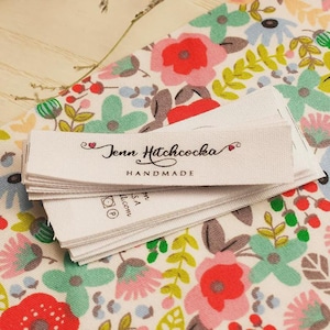 以下が含まれることがあります： 「Jenn Hitchcocka HANDMADE」の黒い筆記体で書かれた白い布ラベルの山。ラベルは、赤、ピンク、青の花柄の生地の上にあります。