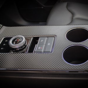 Könnte beinhalten: Nahaufnahme der Mittelkonsole eines Autos mit einer Carbonfaser-Oberfläche. Die Konsole verfügt über einen Gangwahlschalter, zwei Getränkehalter und eine Reihe von Knöpfen.