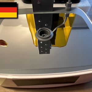 May include: A white, square-shaped device with a reflective, grey surface. The device has a black, circular object on top with a small, black tube extending from it. The device is sitting on a wooden surface.