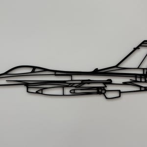 Könnte beinhalten: Ein schwarzes, flaches, zweidimensionales Wandkunstobjekt in Form des Seitenprofils eines Kampfjets. Das Design besteht aus dünnen, miteinander verbundenen Linien, die die Silhouette des Flugzeugs bilden, einschließlich Cockpit, Flügel und Leitwerk.