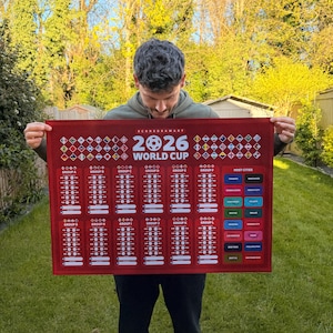 May include: A red World Cup 2026 wall hanging featuring a soccer ball graphic and the text "2026 World Cup". The design includes team flags, group listings, and host cities. The wall hanging is held up by a person.