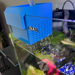 May include: A blue aquarium filter, branded "AQUAEL", is positioned within a fish tank. Water is being poured into the filter. The filter features a perforated front and is partially submerged. The aquarium contains aquatic plants and decorative features.