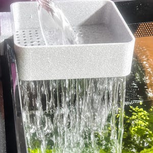 May include: A grey aquarium filter with water pouring into a tank. The filter is square with a textured surface and small holes. The water flows over green aquatic plants.