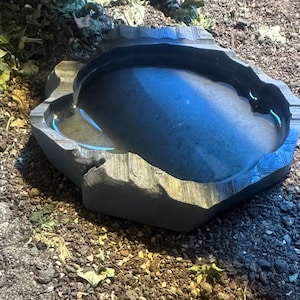 May include: A dark grey, stone-like water dish with a textured, irregular edge. The dish is filled with water, reflecting the sky. The dish is designed for reptiles or small animals, and sits on a bed of soil and small rocks.