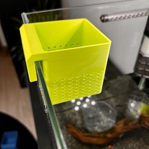May include: A vibrant lime green aquarium feeding cup. The square cup features small holes along the base and side. It is designed to attach to the edge of the aquarium glass. The cup is made from a 3D-printed material.