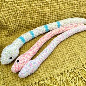Op de afbeelding: Drie gehaakte slangen speelgoed in verschillende kleuren. De eerste slang is wit met blauwe en groene strepen. De tweede slang is roze met witte en gele strepen. De derde slang is wit met roze, blauwe en gele strepen.