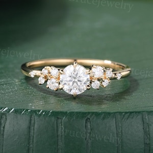 May include: A gold engagement ring featuring a large, round, clear central gemstone. Smaller clear stones are clustered on either side. The ring is set on a dark green surface.
